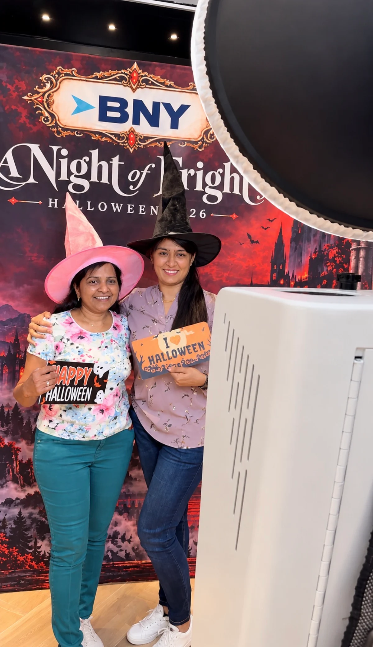 Two guests in witch hats holding Halloween props in front of the BNY custom backdrop